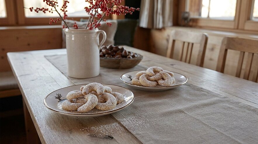 Zwei Teller mit puderzuckerbedeckten Vanillekipferl auf einem rustikalen Holztisch im warmen Tageslicht, daneben Schale mit Maroni und Keramikkanne mit Zweigen.
