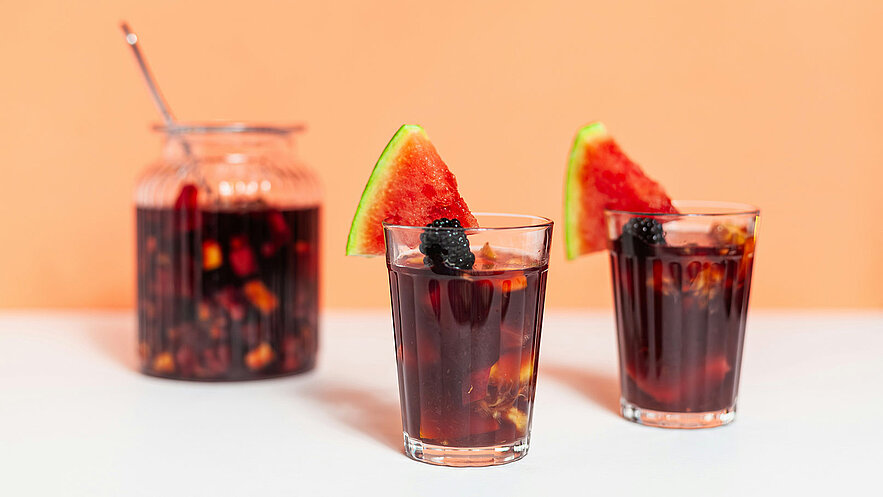 Zwei Gläser mit dunklem Getränk, garniert mit einem Stück Wassermelone und einer Brombeere. Im Hintergrund steht ein großer, ebenfalls gefüllter Glasbehälter mit dem gleichen Getränk. Der Hintergrund ist in sanften Pfirsichtönen gehalten.