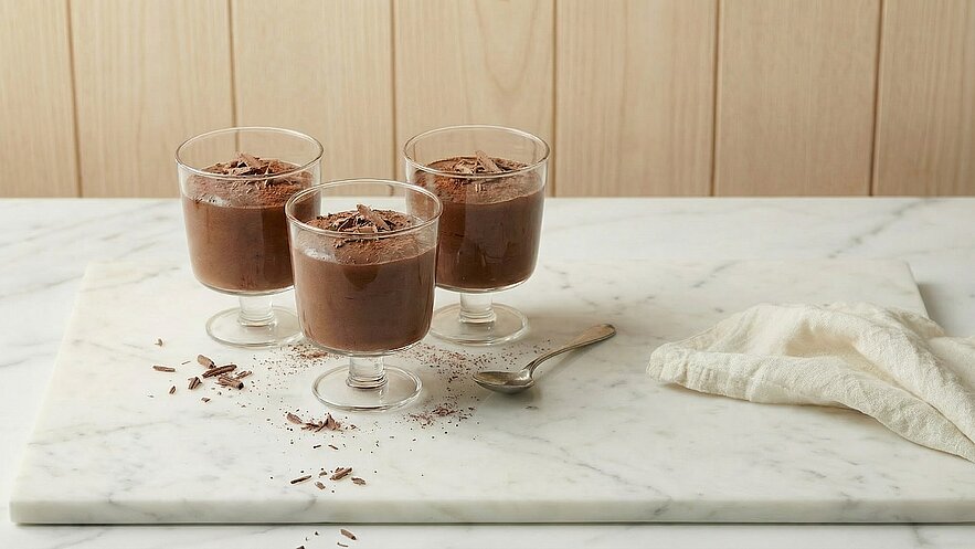 Drei Gläser mit Schokoladenmousse stehen auf einer marmorierten Oberfläche. Die Mousse ist mit Schokoladenraspeln garniert. Neben den Gläsern liegt ein Löffel und ein weißes Tuch.