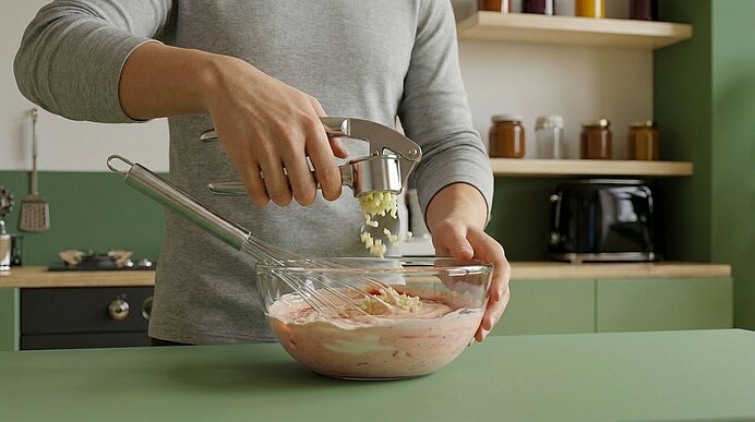 Oberkörper einer Person, die mit einer Knoblauchpresse gehackten Knoblauch über einer Glasschüssel mit rosa Marinade presst; in der Schüssel liegt ein Schneebesen.