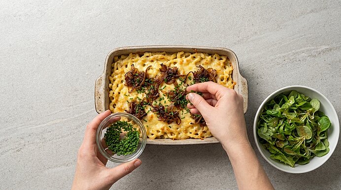 Hände streuen gehackten Schnittlauch auf einen frisch gebackenen Nudel-Käseauflauf mit karamellisierten Zwiebeln; daneben eine Schüssel grüner Salat.