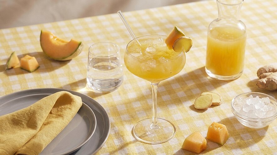 Auf kariertem gelben Tischtuch: ein gekühlter Melonencocktail in einer Coupette mit Melonenscheibe und Strohhalm, daneben Karaffe mit Saft, Glas Wasser, Melonenstücke, Ingwerscheiben und Eiswürfel.