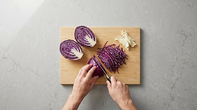Hände schneiden auf einem Holzbrett lila Rotkohl; zwei Kohlhälften und ein Haufen fein geschnittener Streifen sichtbar.