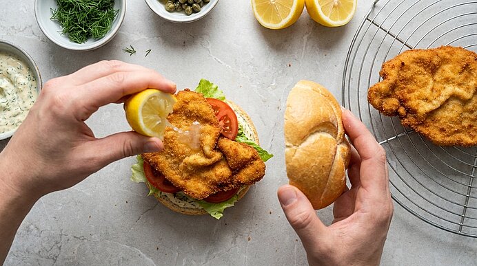 Eine Hand träufelt Zitrone auf das Schnitzel im belegten Brötchen, die andere hält die obere Brötchenhälfte.