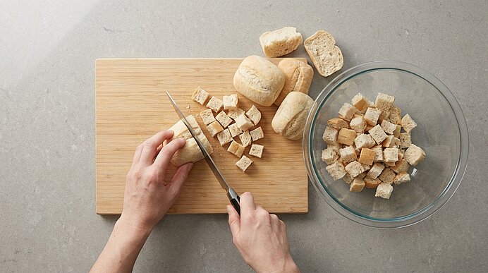 Zwei Hände schneiden mit einem Messer Brötchen in Würfel auf einem Holzbrett; eine Glasschüssel mit bereits gewürfeltem Brot steht daneben.
