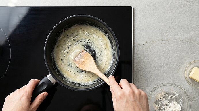 Hände rühren mit Holzlöffel eine schäumende Butter- und Mehlsauce in einem kleinen Topf auf dem Ceranfeld; Schälchen mit Butter und Mehl stehen daneben.