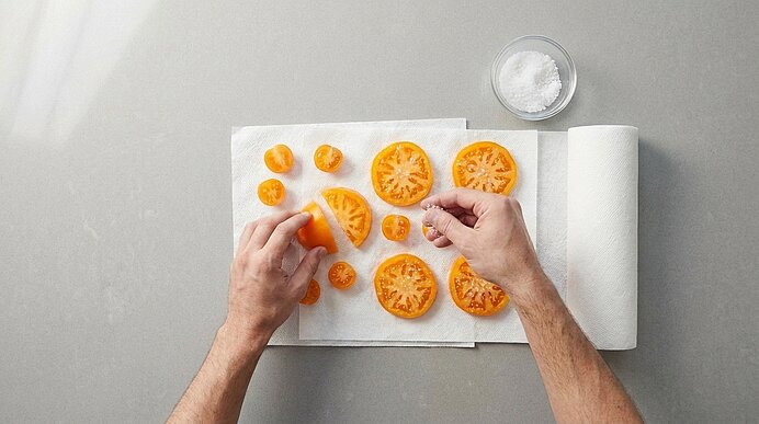 Hände legen gelbe Tomatenscheiben auf ein Blatt Küchenpapier; daneben eine kleine Schale Salz und eine Küchenrolle.