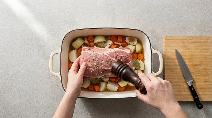 Zwei Hände würzen mit einer Pfeffermühle einen rohen Schweinebraten, der auf geschnittenem Gemüse in einem Bräter liegt.