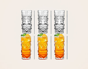 Sechs farbige Trinkgläser mit Tiki-Muster sind abgebildet. Oben stehen zwei leere Gläser, darunter vier gefüllte Gläser mit einem orangefarbenen Getränk, verziert mit einem Minzblatt und einer Zitronenscheibe. Die Gläser sind auf einem hellen Hintergrund angeordnet.