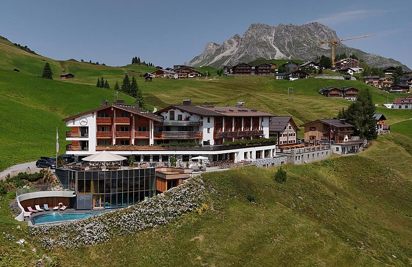 Ein modernes Hotel in einer hügeligen Alpenlandschaft. Im Vordergrund ist ein Poolbereich sichtbar, umgeben von Blumen. Im Hintergrund sind weitere alpine Hütten und Berge, die eine malerische Kulisse bieten. Der Himmel ist klar und die Umgebung wirkt einladend und naturnah.
