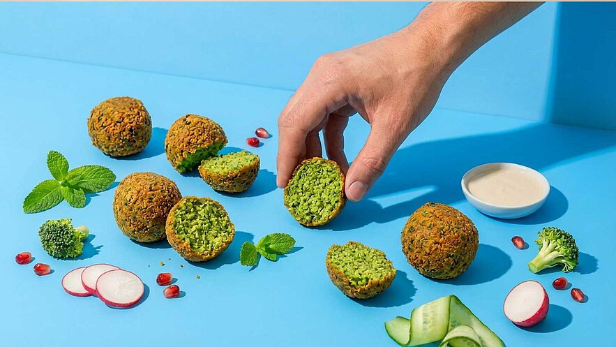 Eine Hand greift nach einem Kichererbsen-Bällchen, das auf einem blauen Hintergrund liegt. Um die Bällchen herum sind geschnittene Zutaten wie Radieschen, Gurken und Brokkoli sowie ein kleiner Dip in einer Schale angeordnet. Zwei Bällchen sind aufgeschnitten, revealing die grüne Füllung.