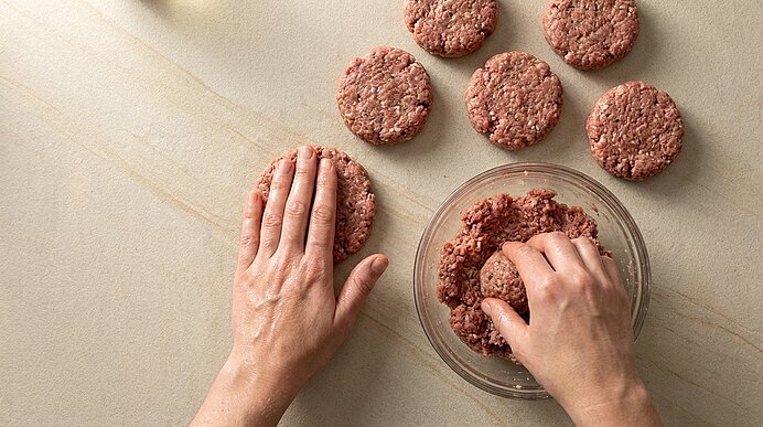 Hände formen rohe Hackfleisch-Patties aus einer Glasschüssel; mehrere bereits geformte Fleischfrikadellen liegen auf der Arbeitsfläche.