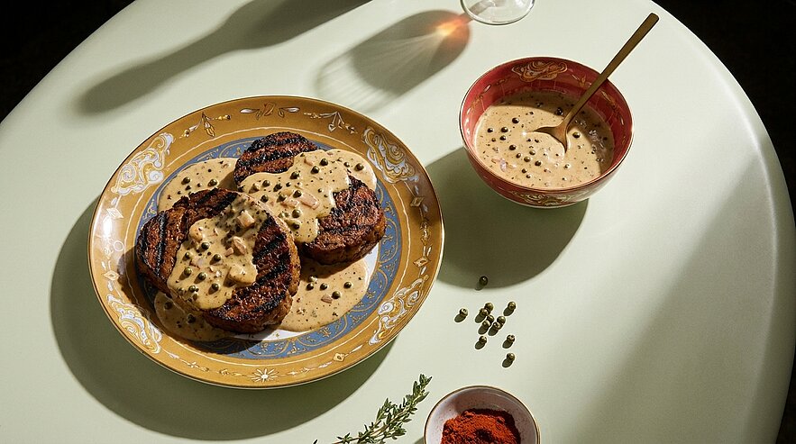 Auf dem Tisch liegt ein farbenfrohes, rundes Teller mit drei gegrillten Patties, die mit einer cremigen Sauce bedeckt sind. Neben dem Teller steht eine rote Schüssel mit einer zusätzlichen Sauce und einem Löffel. Einige Gewürze sind um die Schüssel verteilt.