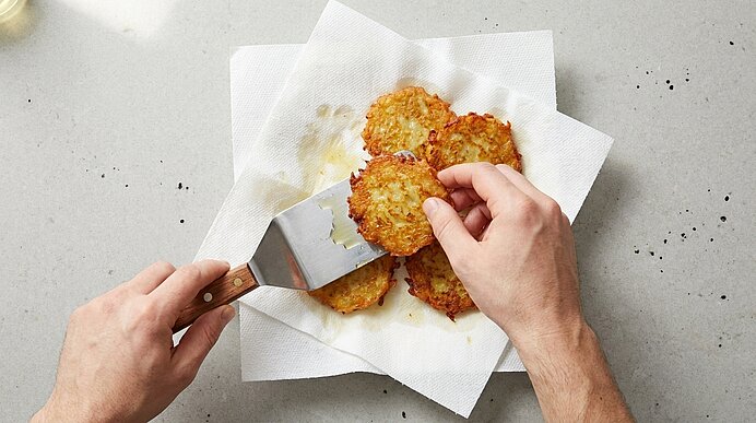 Zwei Hände legen mit einem Pfannenwender goldbraune Kartoffelpuffer auf Küchenpapier zum Abtropfen von Fett.
