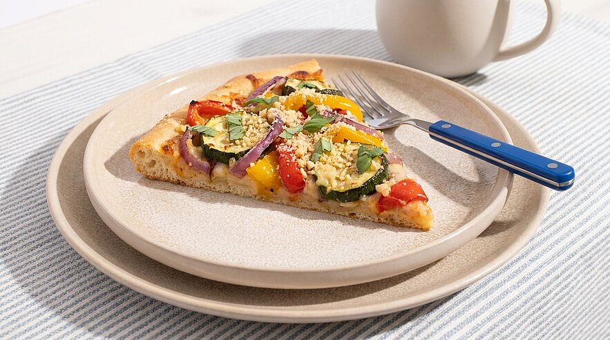 Scheibe vegetarischer Pizza mit Tomaten, gelber Paprika, Spinat, roten Zwiebeln und zerbröseltem Käse auf einem hellen Teller, daneben Gabel und Becher.