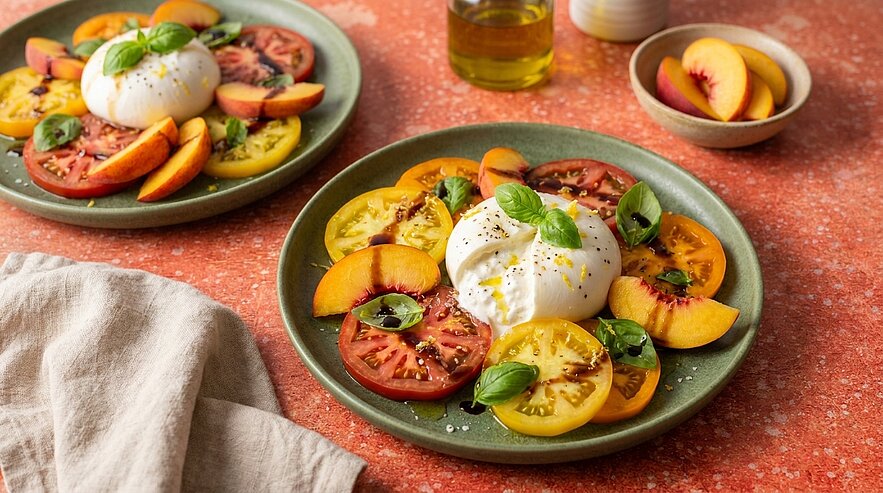 Burrata-Käse auf grüner Platte, umgeben von bunten Tomatenscheiben und Pfirsichspalten, garniert mit frischem Basilikum und Olivenöl.