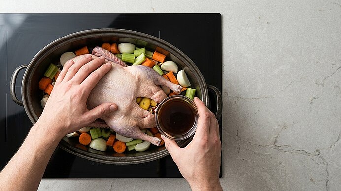 Hände legen ein rohes, ganzes Hähnchen in einen Bräter auf geschnittenes Gemüse (Karotten, Sellerie, Zwiebeln) und gießen Brühe oder Marinade in die Hähnchenhöhle.