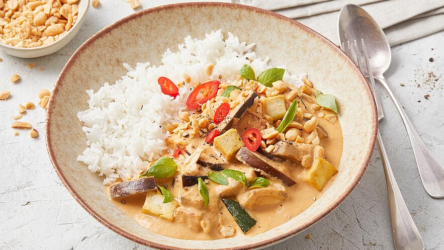 Eine Schale mit aromatischem Curry, das mit Reis serviert wird. Das Gericht enthält Stücke von Gemüse und Fleisch, garniert mit Erdnüssen, roten Chilischoten und frischen Basilikumblättern. Im Hintergrund sind einige Erdnüsse sichtbar.