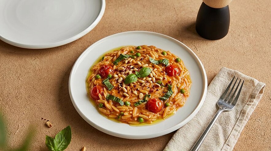 Ein Teller mit Risotto, garniert mit frischem Basilikum, Kirschtomaten und Pinienkernen. Der Hintergrund besteht aus einer neutralen Oberfläche, auf der sich ein Besteckset und ein leeres Geschirr befinden.
