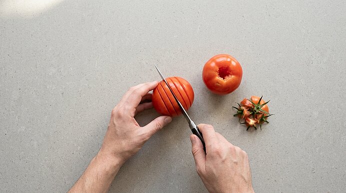Zwei Hände schneiden eine Tomate auf einer hellgrauen Arbeitsplatte mit einem Messer fächerartig ein.