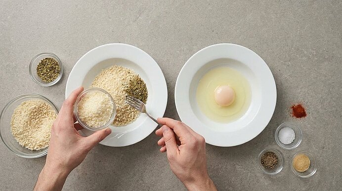 Zwei Hände geben geriebenen Parmesan zu Panko auf einen Teller. Daneben steht ein Teller mit einem rohen Ei.