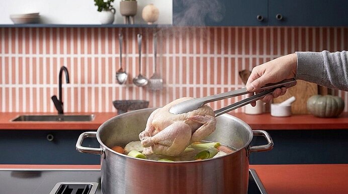 Hand hält mit Zange ein ganzes Huhn über einen dampfenden Topf mit Gemüse auf dem Herd in einer modernen Küche.
