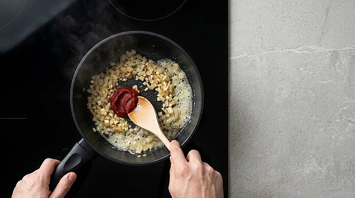 Zwei Hände rühren Tomatenmark mit einem Holzlöffel unter die glasigen Zwiebeln und Knoblauch im schwarzen Topf.