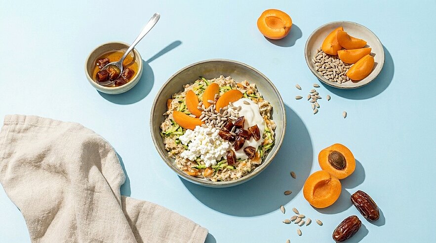 Schüssel mit Haferbrei oder Müsli, garniert mit Aprikosen, Hüttenkäse, Joghurt, gehackten Datteln und Sonnenblumenkernen; daneben frische Aprikosenhälften und Datteln.