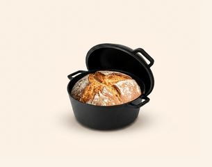 Ein gusseiserner Topf mit frisch gebackenem Brot, das eine goldene Kruste hat, steht im Vordergrund. Im Hintergrund sind eine Schachtel mit Rezepten sowie einige Weizenkörner zu sehen. Der Fokus liegt auf dem Brot und dem Topf.