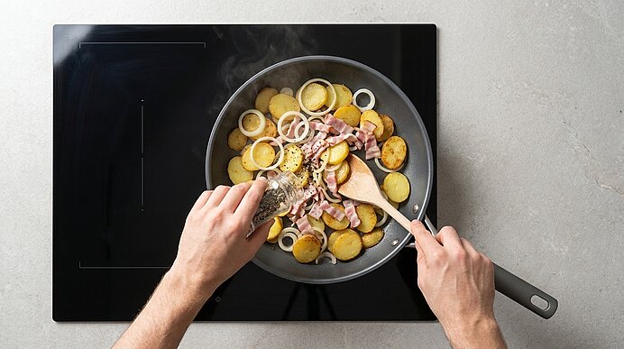 Zwei Hände würzen und rühren in einer Pfanne mit gebratenen Kartoffelscheiben, Zwiebelringen und Speck auf einem Herd.