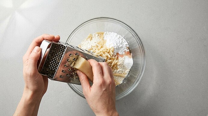 Nahaufnahme: Zwei Hände reiben Butter über eine Vierkantreibe in eine Glasschüssel mit Mehl und weiteren Backzutaten zur Teigzubereitung.