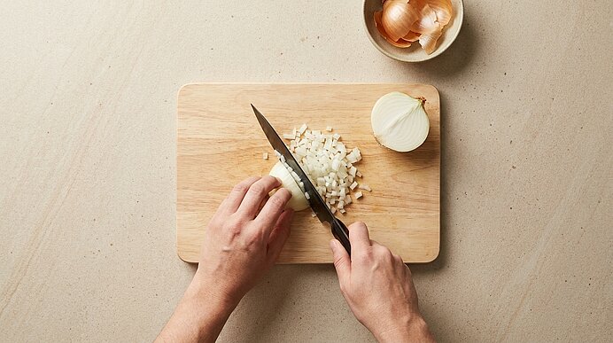 Hände schneiden eine Zwiebel auf einem Holzbrett mit einem Messer; fein gewürfelte Zwiebelstücke liegen daneben.