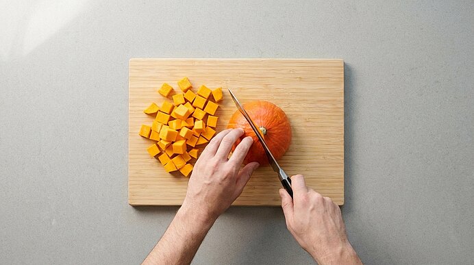 Zwei Hände schneiden einen orangefarbenen Kürbis auf einem Holzbrett mit einem Messer. Daneben liegen Kürbiswürfel.