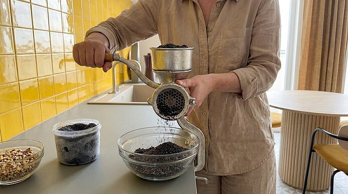 Eine Person mahlt Kaffeebohnen mit einer handbetriebenen Metall-Kaffeemühle über einer Glasschüssel; frisch gemahlener Kaffee fällt in die Schüssel.
