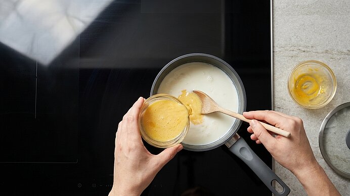 Hände gießen eine gelbe Sauce aus einem Glas in einen Topf mit weißer Flüssigkeit und rühren mit einem Holzlöffel auf dem Herd.
