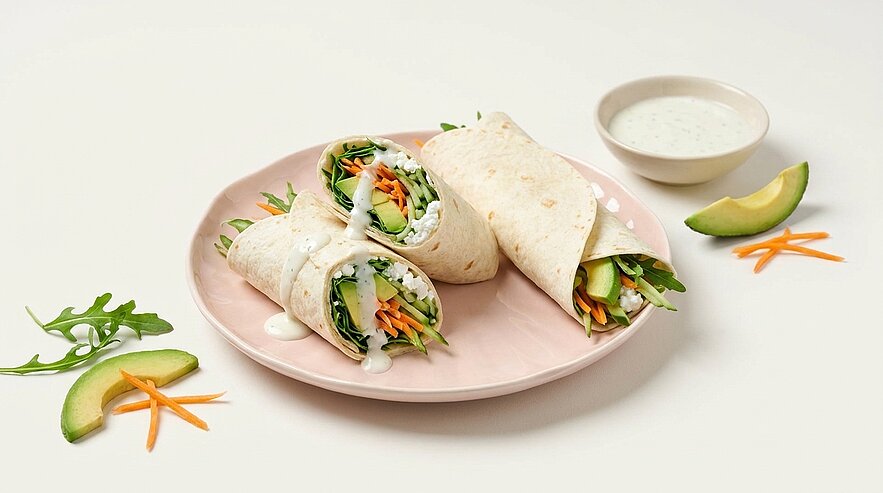 Drei vegetarische Tortilla-Wraps auf einem Teller, gefüllt mit Rucola, Gurke, Karotte und Avocado, dazu ein Schälchen cremiges Dressing.