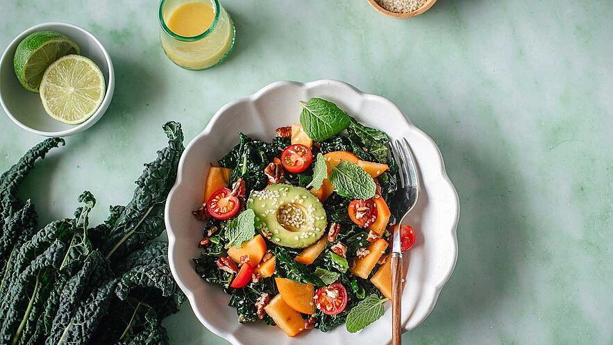 Eine Schüssel mit buntem Salat, bestehend aus frischem Blattgemüse, Kirschtomaten und orangefarbenen Stücken, möglicherweise von einer Melone oder einer ähnlichen Frucht. Ein Essbesteck liegt daneben, während einige Zutaten im Hintergrund sichtbar sind.