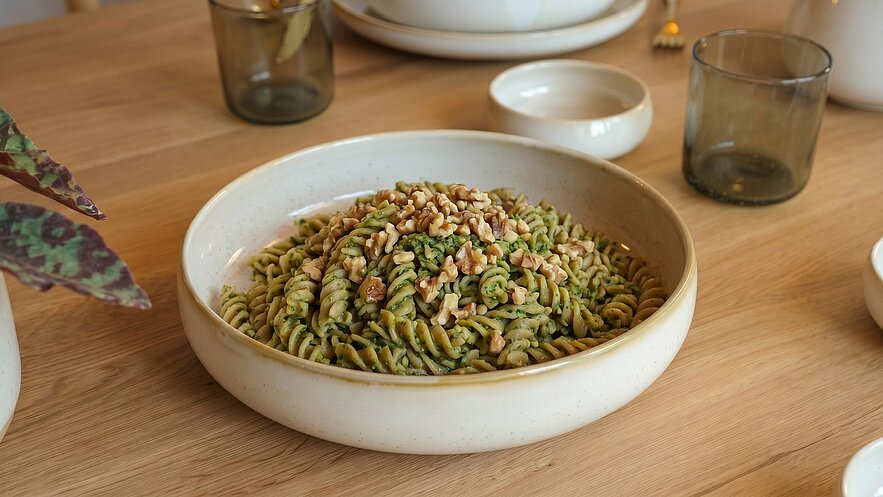 Eine flache Schüssel mit grünen Fusilli-Pasta, garniert mit gerösteten Pinienkernen. Der Hintergrund zeigt einen Holztisch mit weiteren Geschirrstücken und einem Pflanzenblatt.