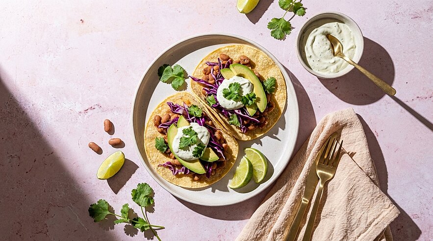 Zwei vegetarische Maistacos auf einem Teller, gefüllt mit Bohnen, Rotkohl, Avocadoscheiben, einem Klecks Sauerrahm und frischem Koriander, dazu Limettenspalten.