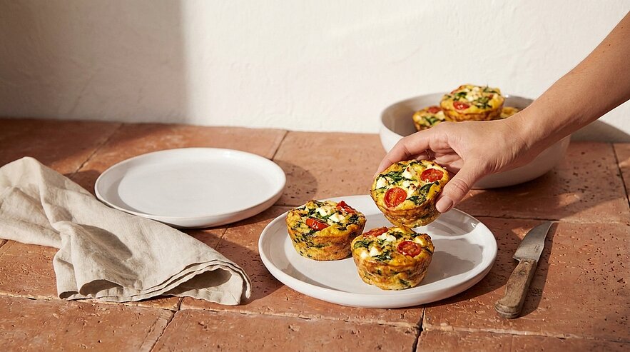 Hand nimmt einen herzhaften Eier‑Muffin mit Cherrytomaten, Spinat und Kräutern von einem weißen Teller; zwei weitere Muffins liegen auf dem Teller.