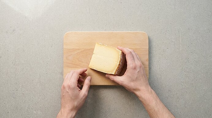 Zwei Hände platzieren ein Stück Hartkäse mit Rinde auf einem kleinen Holzbrett auf einer Küchenarbeitsplatte.