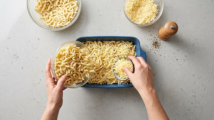 Hände geben gekochte Pasta in eine Auflaufform und bestreuen sie mit geriebenem Käse; zwei Schalen mit zusätzlicher Pasta und Käse stehen daneben.