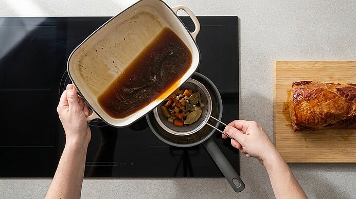 Hände gießen Bratensaft aus einem weißen Bräter durch ein Sieb in einen kleinen Topf auf dem Herd; rechts liegt ein gebratener Braten auf einem Holzbrett.