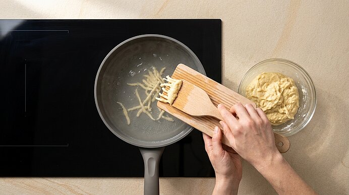 Zwei Hände drücken mit einem Holzbrett und Spatel geriebene Kartoffeln in eine Pfanne auf dem Herd; daneben eine Glasschüssel mit restlicher Kartoffelmasse.