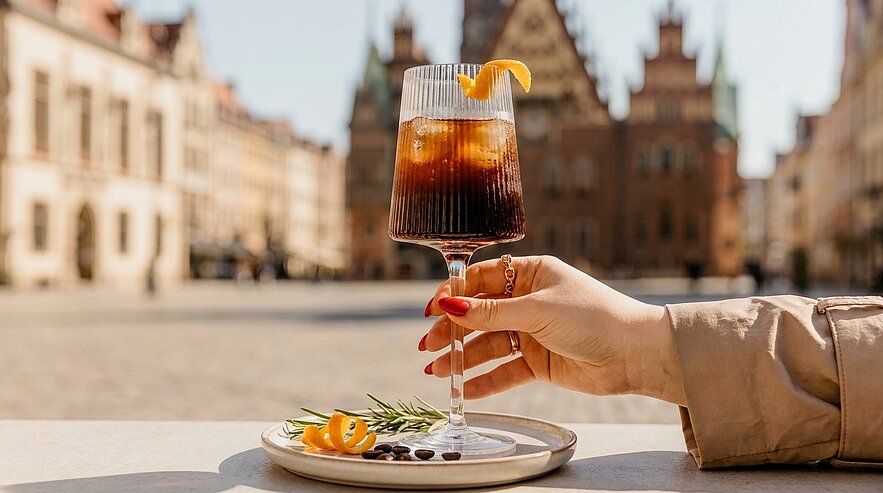 Hand mit rotem Nagellack hält Stielglas mit kühlem Kaffeegetränk, garniert mit Orangenzeste; kleiner Teller mit Kaffeebohnen und Rosmarinzweig auf Tisch.