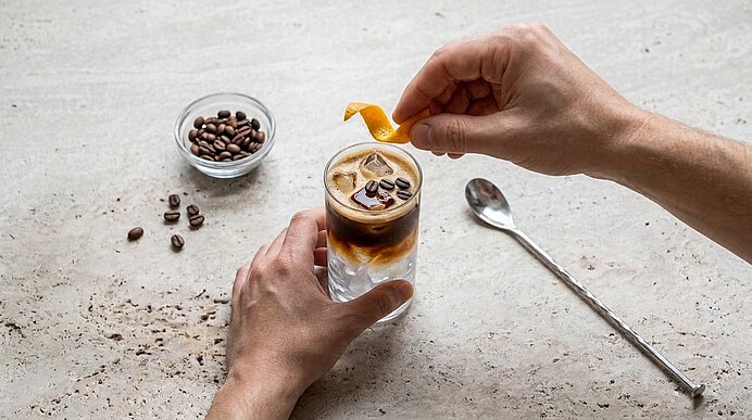 Zwei Hände halten und garnieren ein Glas kalten Espresso mit Eiswürfeln; eine Hand setzt eine Zitronen- oder Orangenzeste als Garnitur.