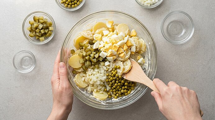 Zwei Hände rühren mit einem Holzlöffel in einer Glasschüssel Kartoffelsalat aus Kartoffelscheiben, Eiern, Erbsen, Gewürzgurken und Zwiebeln.