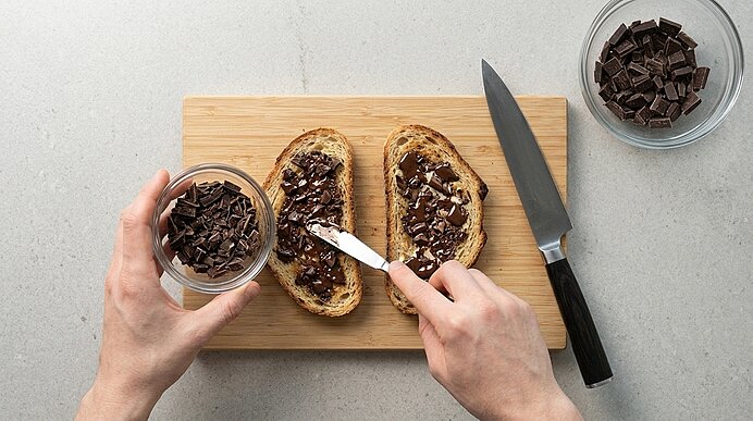 Zwei Hände verstreichen leicht geschmolzene gehackte Schokolade auf gerösteten Brotscheiben auf einem Holzbrett.
