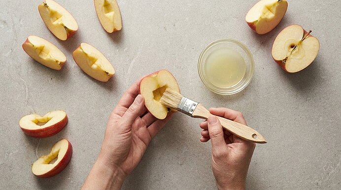 Zwei Hände bestreichen eine ausgehöhlte Apfelspalte mithilfe eines Pinsels mit Zitronensaft aus einer Glasschale.