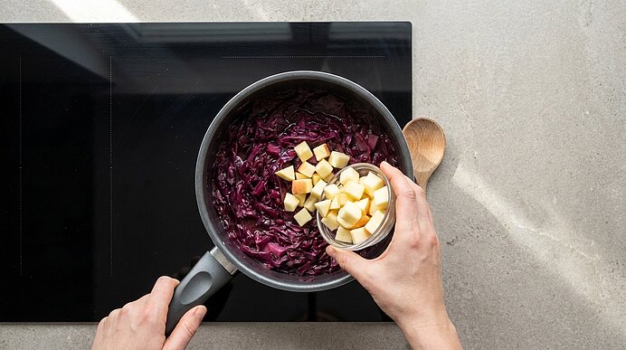 Zwei Hände geben gewürfelte Äpfel in einen Topf mit geschmortem Rotkohl auf dem Herd; daneben liegt ein Holzlöffel.
